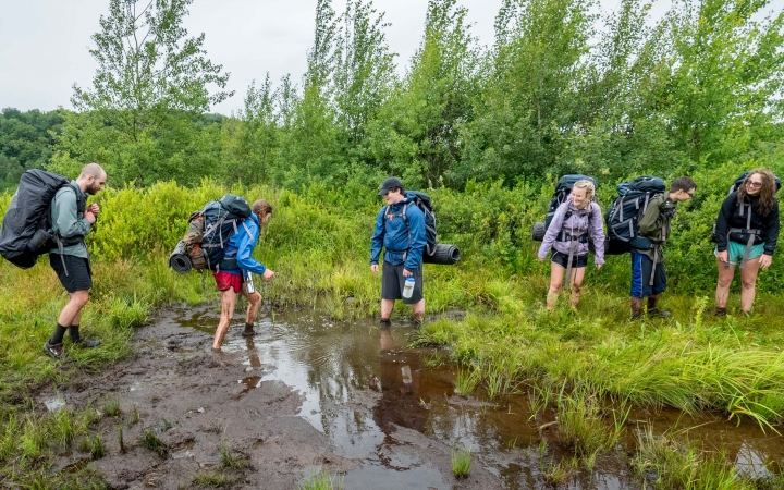 A group of young people wearing backpacks make their way though grass and ankle-deep mud.